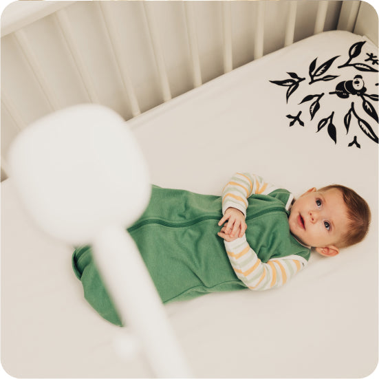 Baby lying on a Nanit Smart Sheet with Spruce Green Sleep Wear Sleeping Bag and with Nanit Pro Baby Monitor with Floor Stand at bird's eye view