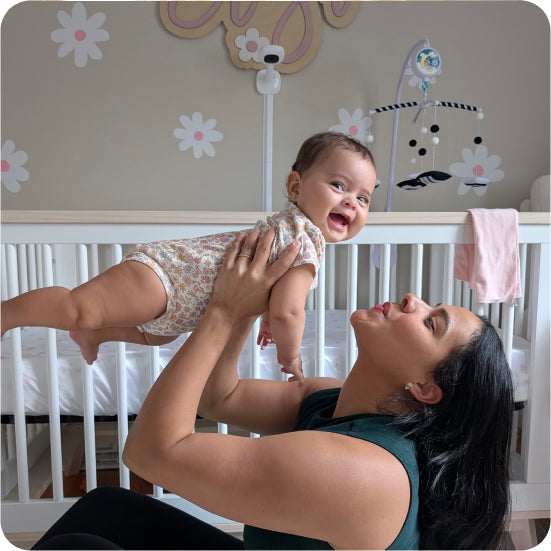 Woman lifting a baby in a crib with decorative wall stickers and Nanit Pro Baby Monitor + Wall Mount in the background